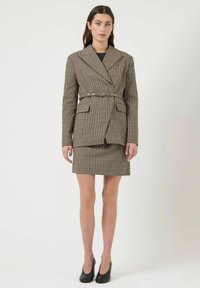 Brun ternet blazer med en markeret talje, kombineret med et matchende skørt. Har skarpe revers og forlommer. Sorte højhælede sko.