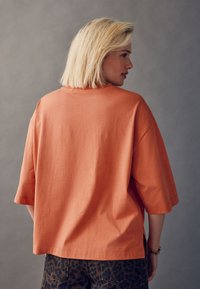 Lõdvalt istuv, lühikeste varrukatega särtsakas oranžist puuvillasest materjalist T-särk. Omaduseks on ümara kaelusega ja küljetagused lõhikud, mida on sobitatud leopardimustriliste pükstega.