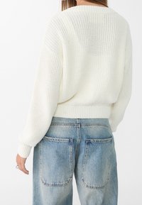 Witte gebreide trui met een geribbelde zoom en manchetten, gecombineerd met lichtblauwe loszittende spijkerbroek met grote achterzakken.