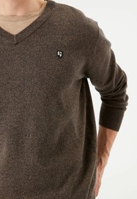 Pull en maille marron avec un col en V et des manches longues. Carpente un motif texturé et un petit logo sur la poitrine gauche.