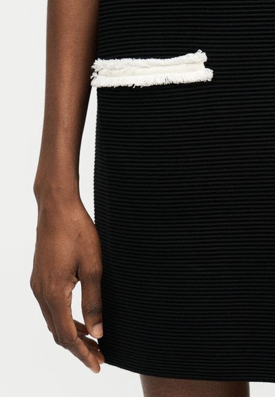 Dunkelhäutige Hand ruht neben schwarzem, strukturiertem Rock mit weißem, fransigem horizontalem Taschendetail auf hellem Hintergrund.