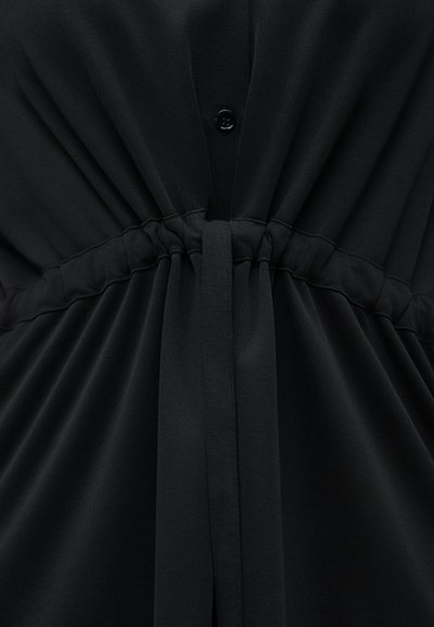 Μαύρο ένδυμα με ελαστική μέση που μαζεύει το ύφασμα και στενή ζώνη από ύφασμα δεμένη μπροστά, με μερικώς εμφανή μαύρο κουμπί πάνω από τη μέση.