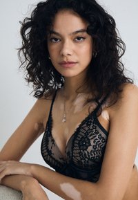 Soutien-gorge en dentelle noire avec des motifs floraux et des bretelles ajustables. Le matériau est transparent avec un bandeau de soutien et des accents matériels discrets.