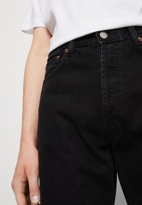 Černé džíny z denimu s vysokým pasem, doplněné měděnými nýty, kovovým zapínáním na knoflík a bočními kapsami. Nosit s bílým tričkem.