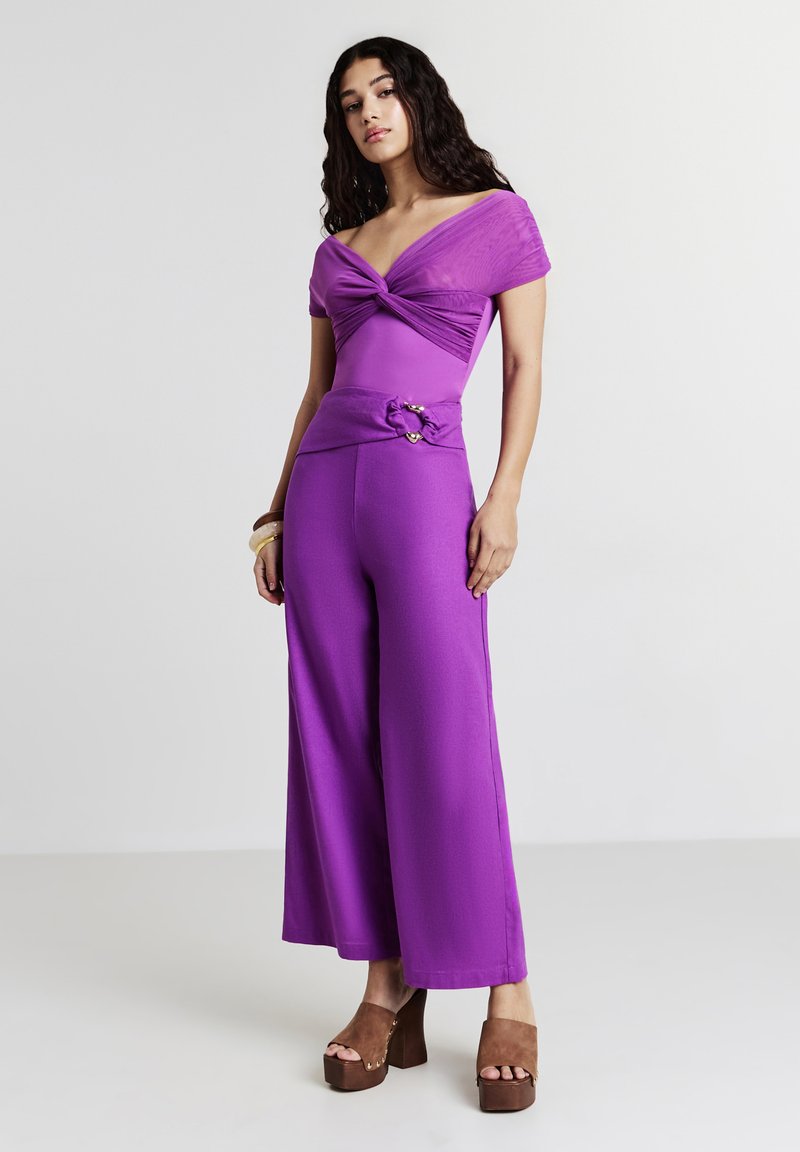 Donna con lunghi capelli ricci che indossa un top viola a spalle scoperte e pantaloni larghi coordinati, in piedi contro uno sfondo grigio uniforme.