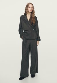 Donkergrijze op maat gemaakte pak met een wikkelstijl blazer, twee voorzakken en brede broekspijpen. De stof heeft een gestructureerde uitstraling.