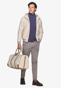 Beige zip-up hoodie, marineblå turtleneck, lysegrå bukser, brune sko, holder en stor beige duffeltaske med brune detaljer og håndtag.