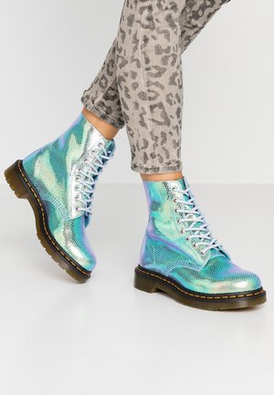 Osoba w błyszczących, opalizujących niebiesko-zielonych butach sznurowanych i szarych spodniach w cętki lamparcie, stojąca na jasnej podłodze na jednolitym tle.