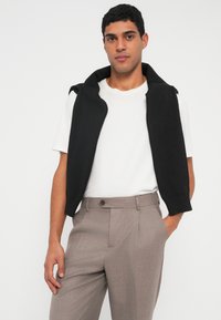 Μαύρο πλεκτό γιλέκο με γιακά φορεμένο πάνω από λευκό μπλουζάκι T-shirt, συνδυασμένο με ανοιχτό καφέ παντελόνι σε ραμμένη γραμμή. Απλός και λειτουργικός σχεδιασμός.