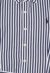 Detailní záběr na košili s námořnickými a bílými vertikálními pruhy, s bílými knoflíky a malým vyšitým logem polo hráče na hrudi.