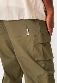 Khaki cargo pants met meerdere zakken, een klein merklabel en een gladde afwerking, gecombineerd met een lichtgekleurde stoffen top.