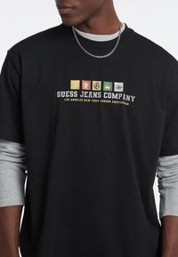 T-shirt nera a maniche corte con il logo della "Guess Jeans Company" e vari design di emblemi colorati; sovrapposta a maniche lunghe grigie visibili.