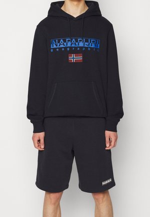 Personne portant un sweat à capuche noir Napapijri avec un logo bleu et un patch du drapeau norvégien, associé à un short noir Napapijri avec un logo blanc.