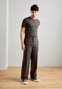 Šedé tričko spárované s šedými proužkovanými kalhotami. Model má na sobě černé tenisky a černou čepici, stojí na dřevěné podlaze.