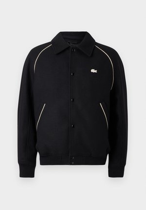 Giacca nera con bottoni a collo, con piping beige lungo le spalle e un piccolo logo ricamato sul lato sinistro del petto.