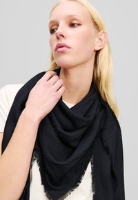 Musta kolmnurkne sall, mis on valmistatud pehmest kangast ja millel on tutid ääres. Peen logo muster üle kogu salli, elegantsetena kaela ümber riputatuna.