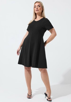 Femme debout, une main sur la hanche, portant une robe noire courte de style trapèze à manches courtes et des sandales noires à talons hauts, sur un fond uni.