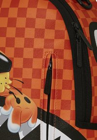 Mochila naranja y negra con un patrón de tablero de ajedrez, gráficos de personajes de dibujos animados y dos bolsillos negros con cremallera con acentos de logo.