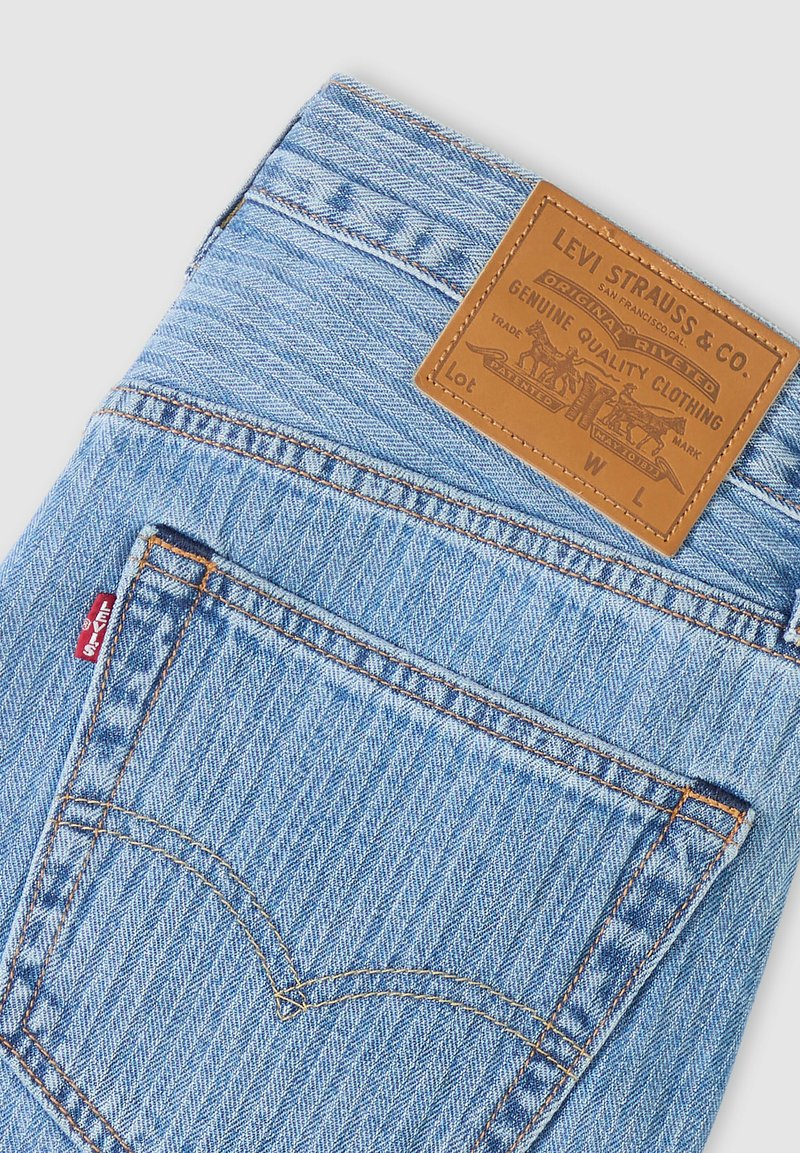 Vaqueros Levi's de mezclilla azul claro que muestran el bolsillo trasero, la etiqueta roja de Levi's y el parche de cuero marrón con el logo y texto de calidad.