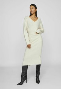 Vestido midi de punto en beige claro con escote en V y mangas largas. Presenta una textura acanalada y una cintura ajustada. Combinado con botas negras hasta la rodilla.