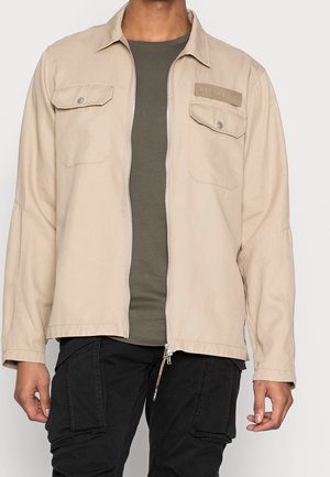 Mand iført beige jakke med lynlås og brystlommer over grøn skjorte og sorte cargobukser, stående foran en ensfarvet baggrund.