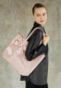 Borsa tote lucida rosa con un grande dettaglio a fiocco, progettata in una forma strutturata, con un logo in rilievo dorato e spallacci spessi.