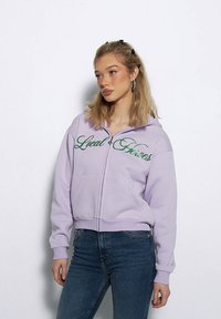 Fioletowa bluza rozpinana z kapturem z napisem "Local Heroes" w zielonym kolorze, o luźnym kroju, z dwoma kieszeniami z przodu, noszona z niebieskimi dżinsami.