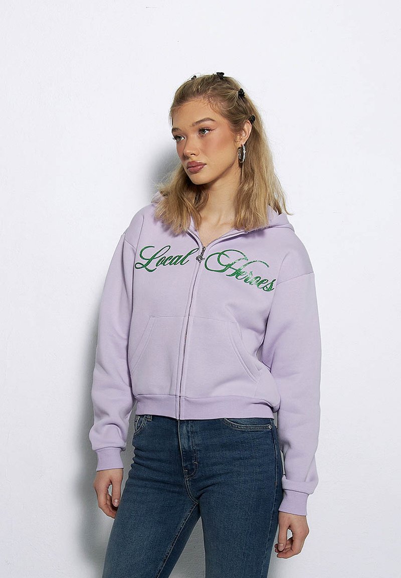 Fioletowa bluza rozpinana z kapturem z napisem "Local Heroes" w zielonym kolorze, o luźnym kroju, z dwoma kieszeniami z przodu, noszona z niebieskimi dżinsami.