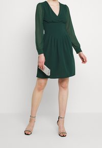 Grön klänning med långa genomskinliga ärmar, djup V-ringning, rynkad midja och utsvängd kjol. Åtföljd av en glittrig silverclutch och svarta klackar.