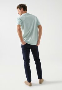 Camiseta de algodón azul claro con mangas cortas, de corte relajado. Combinada con pantalones oscuros de marino y zapatos casuales beige, de pie de manera informal.