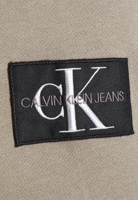 Šedá tkanina s štítkem, na kterém je bílé písmo "CALVIN KLEIN JEANS" na černém pozadí, přišité na tkanině.
