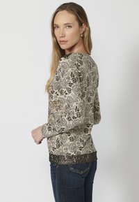 Langarmshirt mit Blumenmuster in Beige und Schwarz, mit einem strukturierten dunkelhäufigen Saum. Weicher Stoff und taillierte Passform.