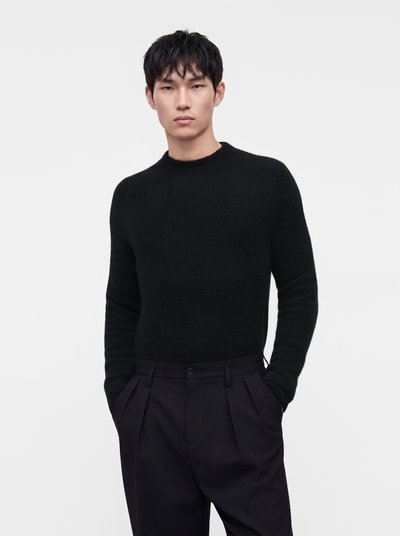 Young man with short black hair wearing a black sweater and black pleated pants with hands in pockets against a plain white background.