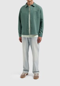 Groen button-up overhemd jack met twee voorzakken, gelaagd over een lichtgroene t-shirt, gecombineerd met lichtblauwe jeans en bruine sneakers.