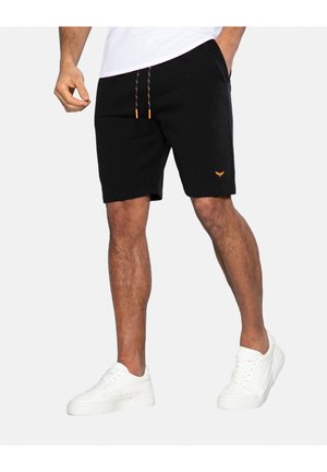 Mann trägt schwarze Shorts mit Kordelzug und kleinem orangefarbenem Logo, weiße Sneaker und weißes T-Shirt, steht vor einem einfarbigen Hintergrund.