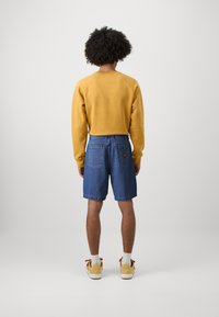 Κίτρινη μπλούζα με ραγκλάν μανίκια, συνδυασμένη με μπλέ τζιν σορτς και ανοιχτόχρωμα υποδήματα. Το φόντο είναι απλό και ουδέτερο.