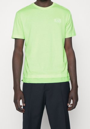 Uomo che indossa una T-shirt a maniche corte di un verde lime brillante con il logo EA7 sul petto e pantaloni neri, in piedi su sfondo bianco.