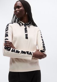 Beige långärmad piké med svartvitt djurmönster på ärmar och krage, med knappgylf och ribbad textur.