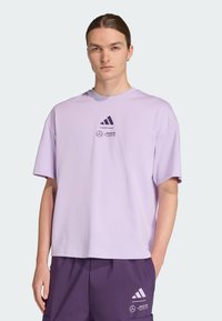 Camiseta oversized de algodón morado claro con cuello redondo, que presenta los logotipos de Adidas y Mercedes en negro en el pecho. Mangas cortas.