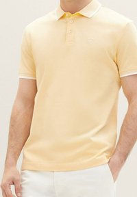 Polo shirt jaune avec une finition texturée, à manches courtes et col rayé blanc. Il dispose d'une patte de boutonnage à trois boutons et d'un petit logo sur la poitrine.