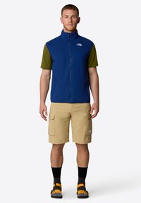 Gilet in pile blu con colletto alto sopra una maglietta verde a maniche corte, abbinato a pantaloni cargo beige, calze nere e sandali arancioni.