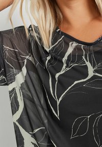 Blusa nera trasparente con motivi a foglia bianchi, tessuto leggero e vestibilità comoda con scollatura arrotondata e maniche corte.