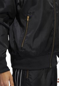 Svart bomberjacka med en glänsande satinfinish, försedd med en guldfärgad dragkedja, elastiska ärmslut och nederkant. Sidofickan med dragkedja ger funktionalitet.