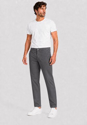 Man draagt een wit t-shirt met korte mouwen, donkergrijze broek en witte sneakers, staat tegen een effen achtergrond en kijkt naar opzij.