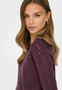 Pull à manches longues de couleur violet profond avec une texture lisse. Présente des paillettes noires et roses sur l'épaule pour un détail supplémentaire.