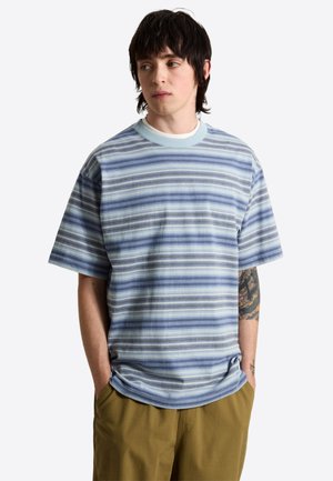 Joven con pelo oscuro, que lleva una camisa de manga corta de rayas azules y grises y pantalones marrones, con las manos en los bolsillos, mirando hacia un lado.