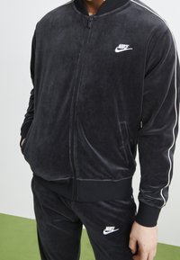 Svart velour zip-up jacka med vita sidostråkar och en Nike-logotyp på bröstet. Har en ståkrage och ribbade cuffs.