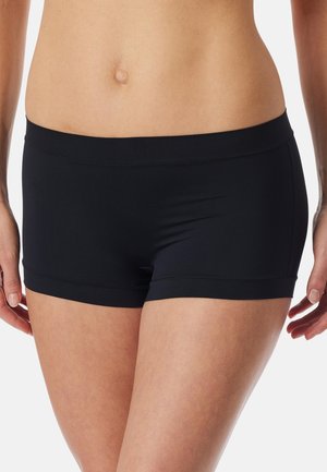Vrouw draagt zwarte, nauwsluitende boxershorts, waarbij de buik en bovenbenen zichtbaar zijn, staand met de armen ontspannen langs het lichaam.