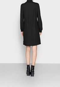 Černé dlouhé šaty s vysokým límcem a volným střihem, vyrobené z texturované látky, končící nad koleny, v kombinaci s černými kotníkovými botami.