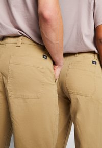Kaki hlače s glatkom teksturom, koje imaju dva stražnja džepa i male oznake brenda. Struk je standardan s petljama za pojas.